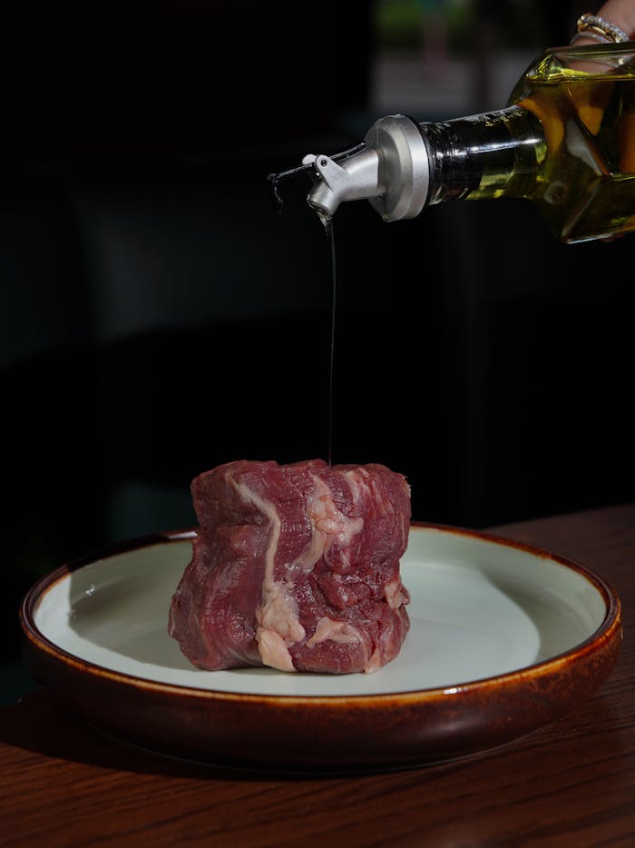 A raw beef steak is drizzled with olive oil on a plate, ready for cooking.